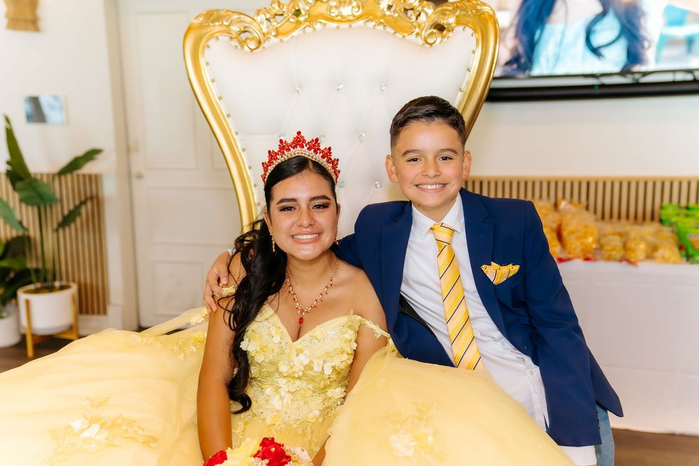 A girl in a yellow quinceañera gown and red crown sits on a gold throne chair with a boy in a navy suit beside her.