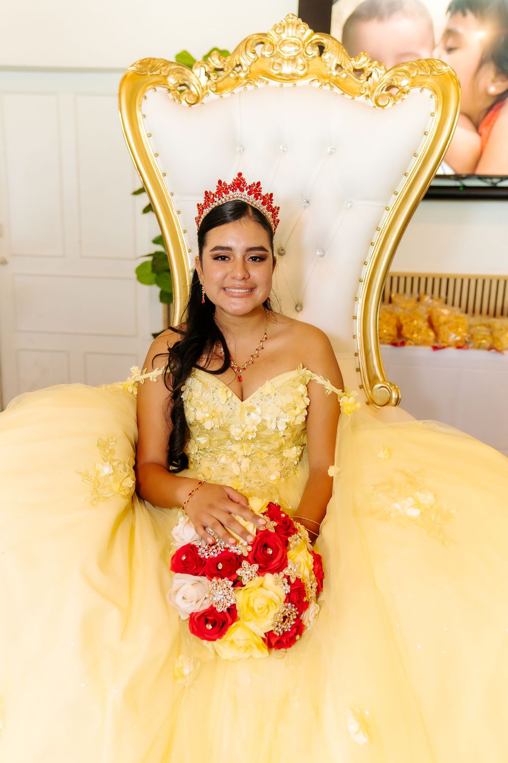 Young woman in yellow quinceañera gown with red tiara sits on ornate gold throne chair holding red and yellow bouquet.