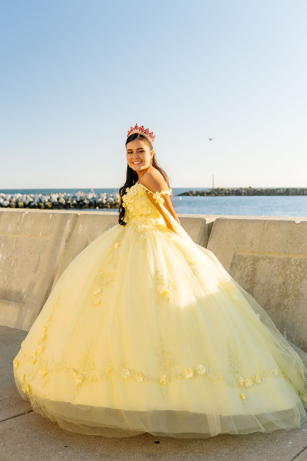 Young woman in yellow ball gown with floral details and red crown poses near waterfront.