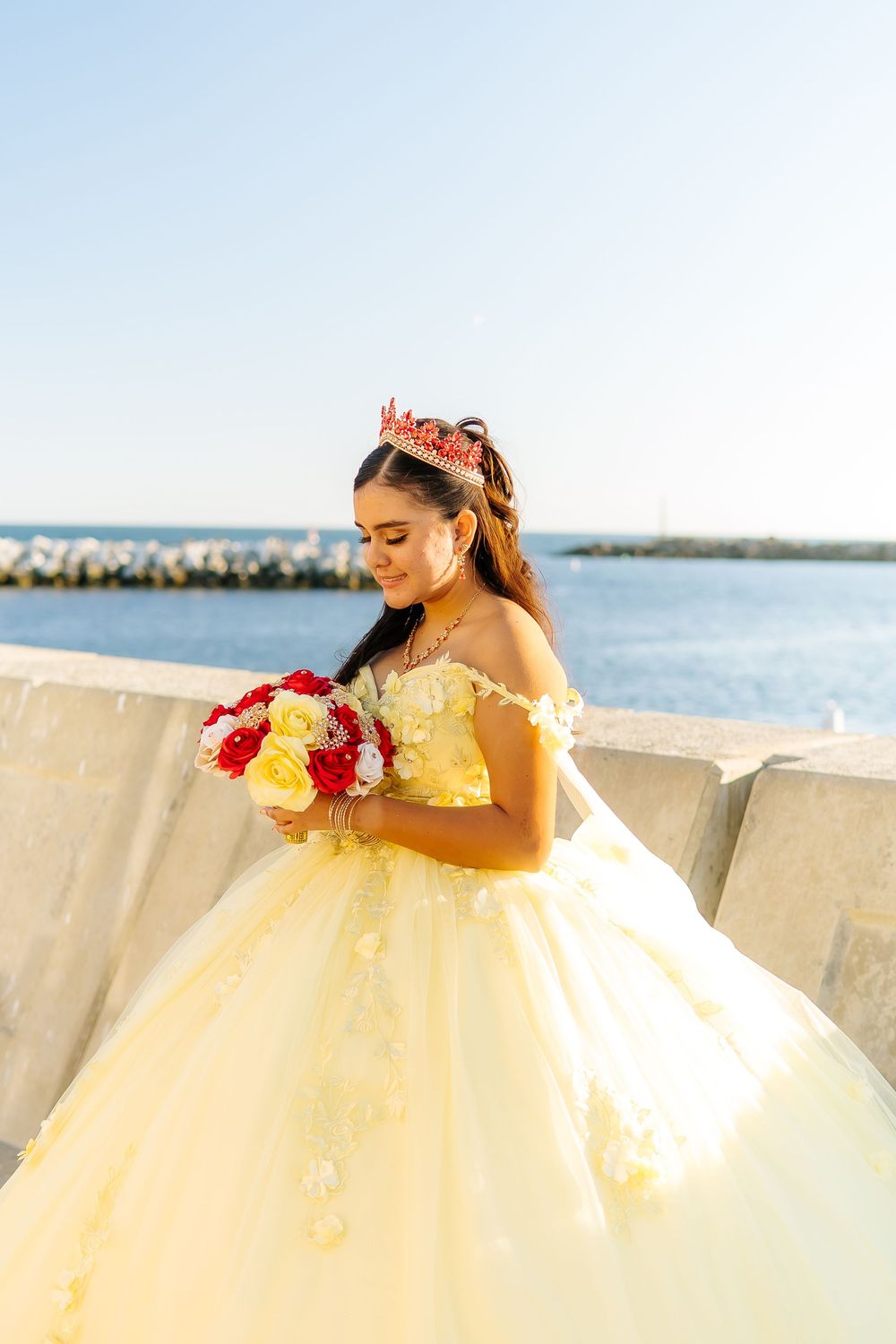 Young woman in yellow quinceañera ball gown with red crown holds bouquet by seaside.