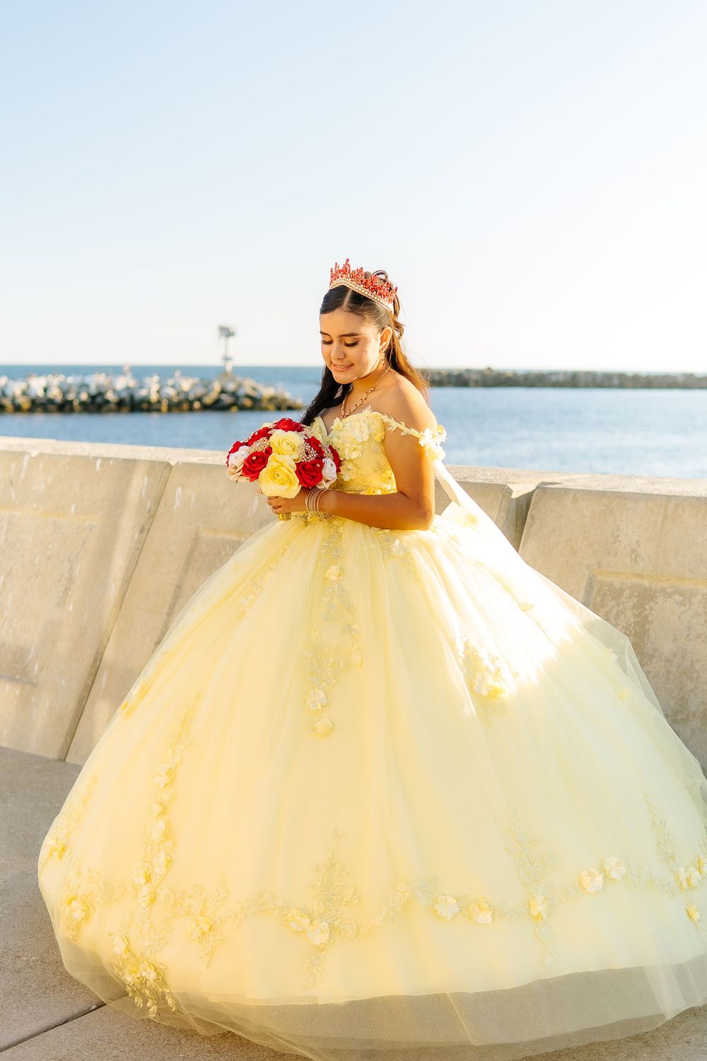 Quinceañera in yellow ball gown holds red and yellow bouquet wearing red tiara by water.