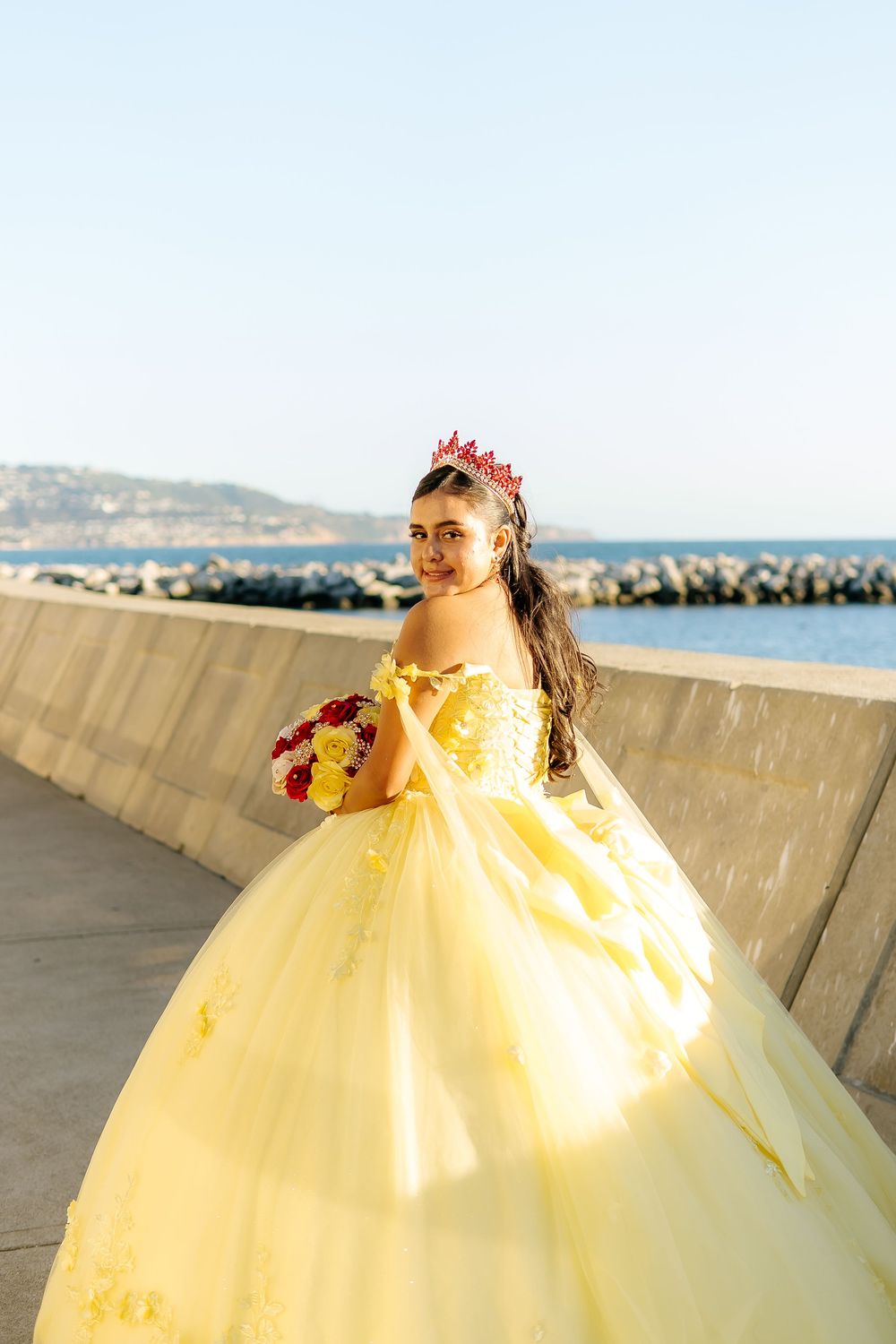 Young woman in yellow ball gown with red crown holds bouquet near ocean for quinceañera celebration.