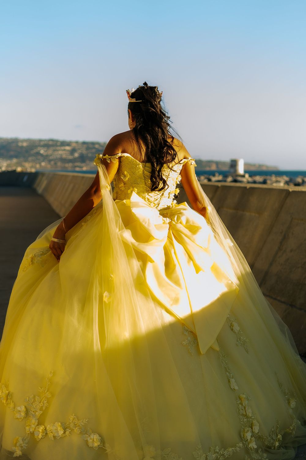 Young woman in elegant yellow quinceañera ball gown with floral details stands outdoors with scenic view behind her.
