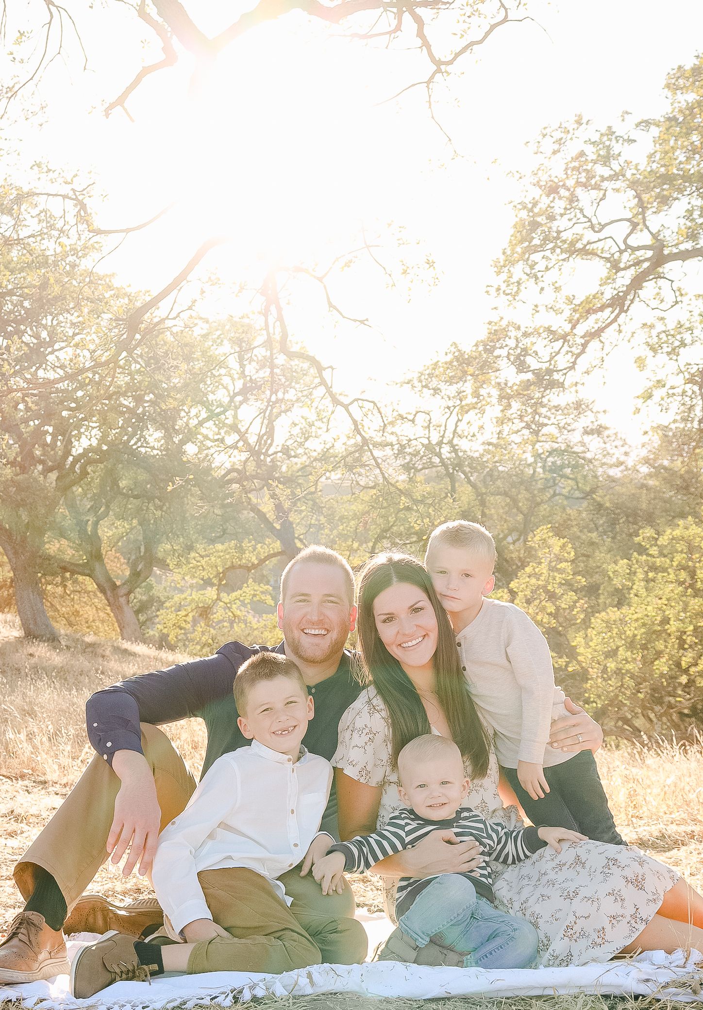 A happy family of five poses outdoors on a blanket surrounded by golden sunlight and oak trees.