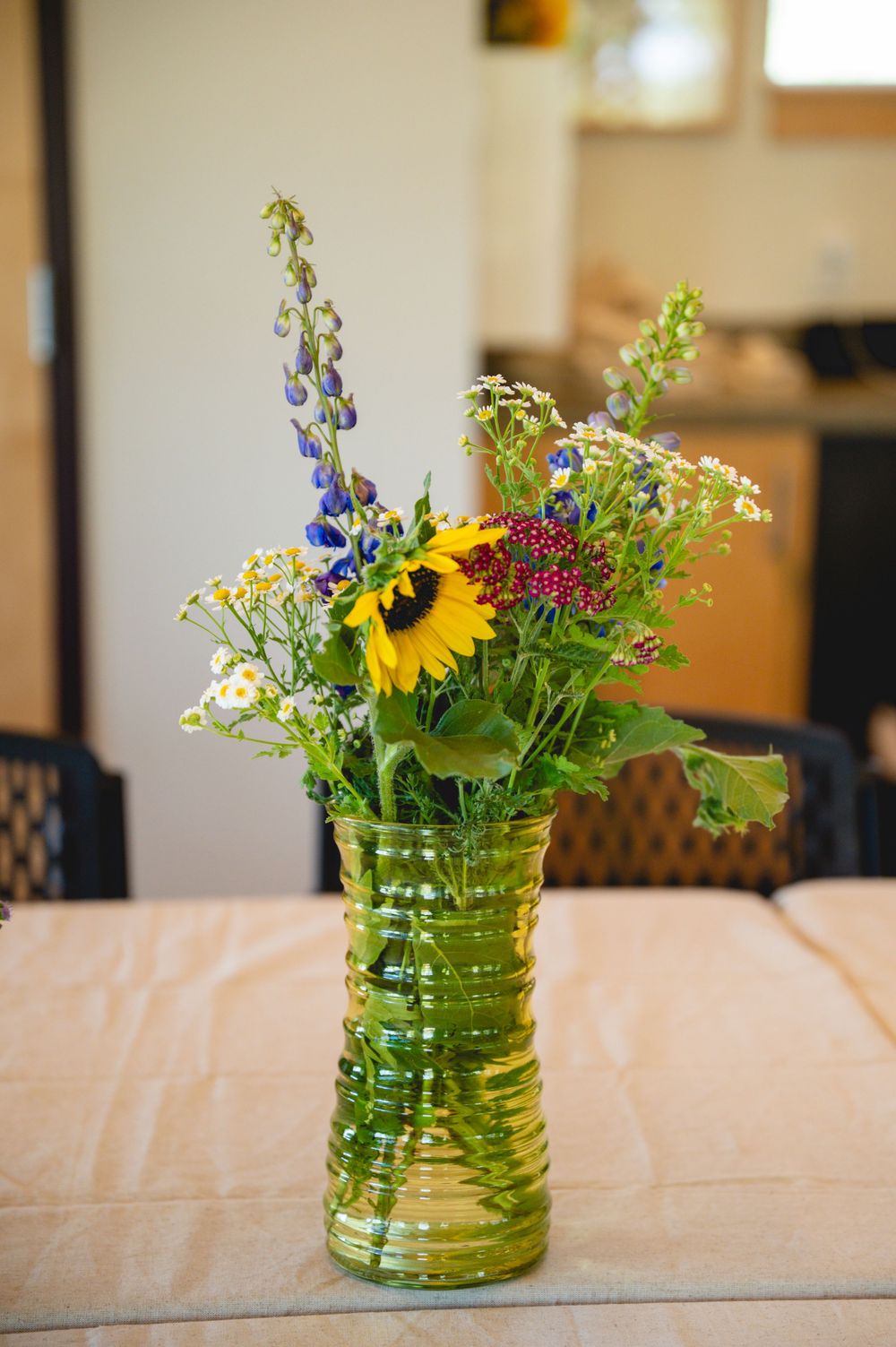 Green ribbed vase with sunflower, blue delphiniums, daisies, and wildflowers as centerpiece on a table indoors.