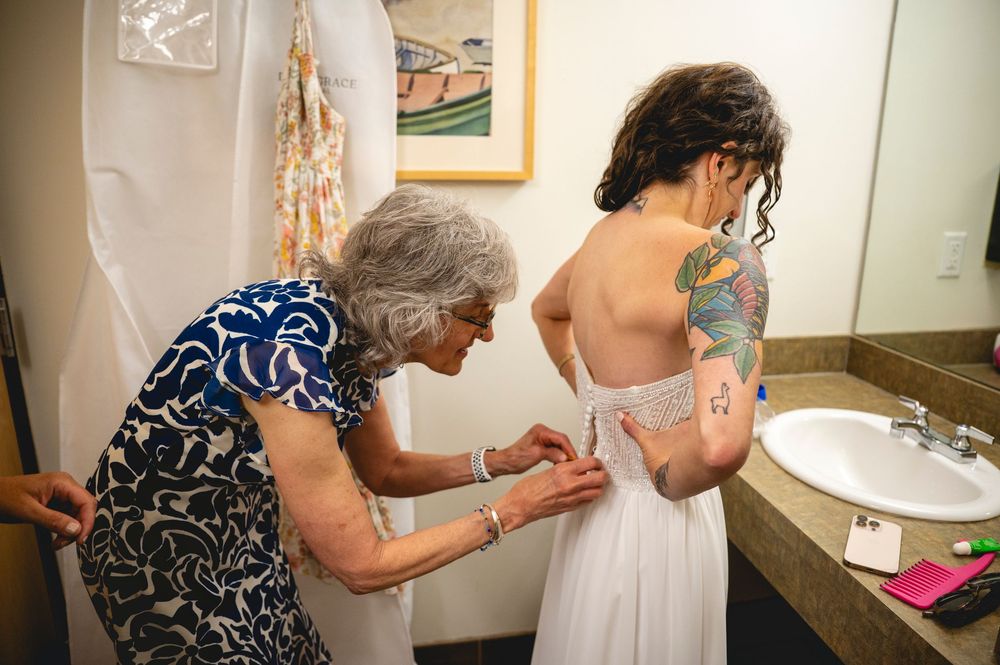 Elderly woman helping a tattooed bride button up her white wedding dress in a hotel bathroom.