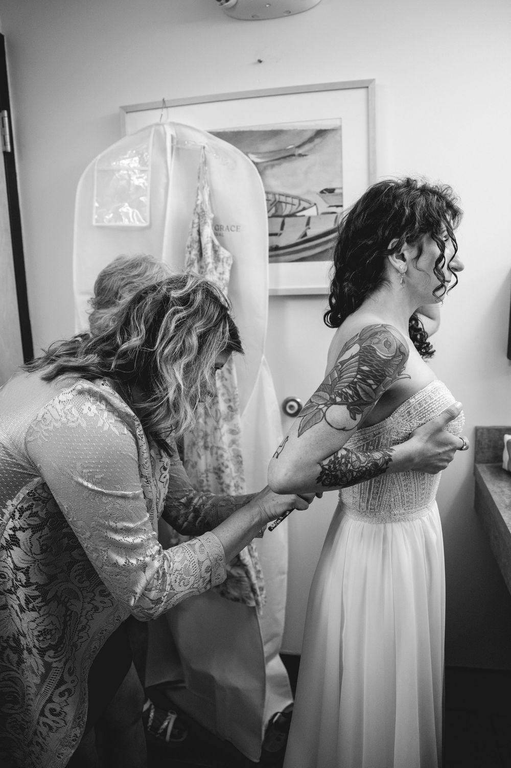 Black and white photo of a bride with tattoos getting her wedding dress fastened by a woman in a lace gown.