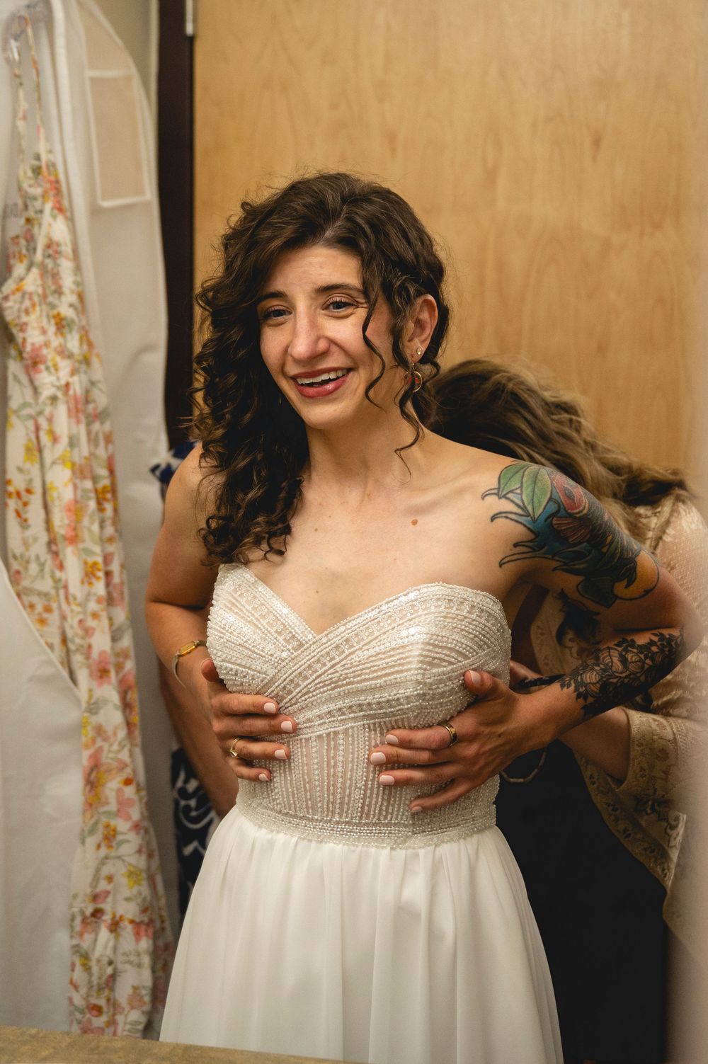 Smiling bride in strapless white lace wedding gown being fitted in a dressing room by a tattooed assistant.