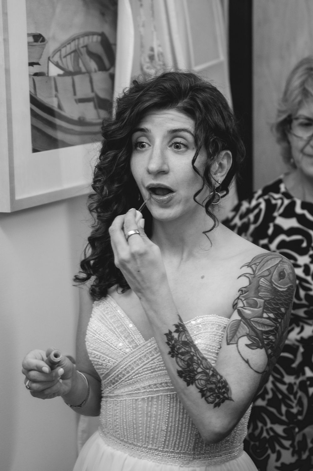 Tattooed bride in white gown applies lipstick, captured in black and white wedding photo.