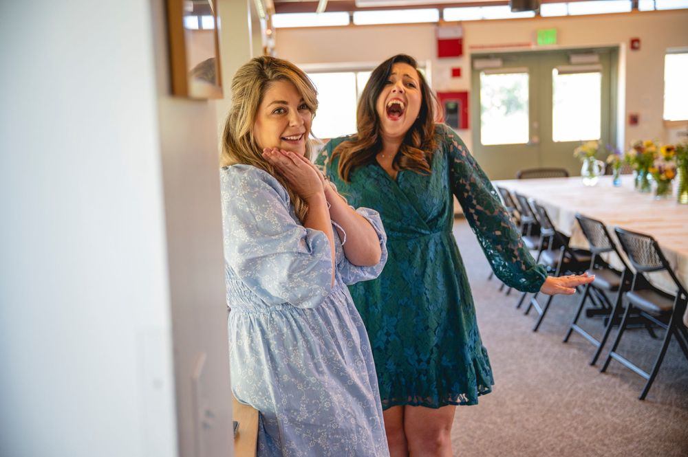 Two women in floral dresses laughing and reacting with surprise in a decorated event hall.