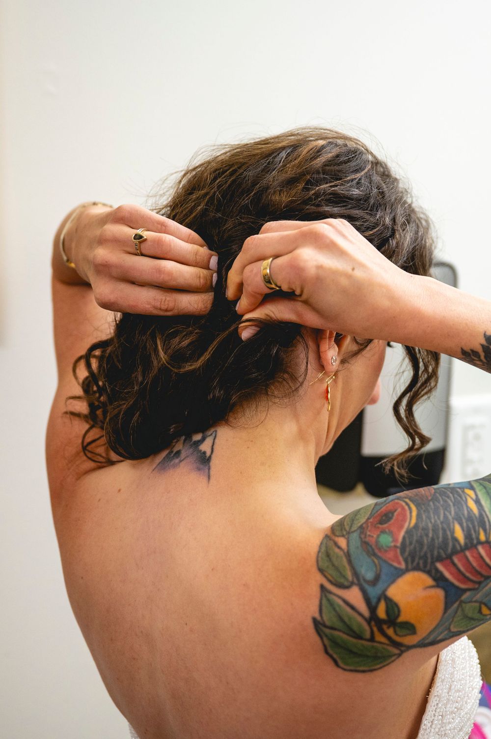 Bride having her curly hair styled up by a tattooed bridesmaid before her wedding ceremony.