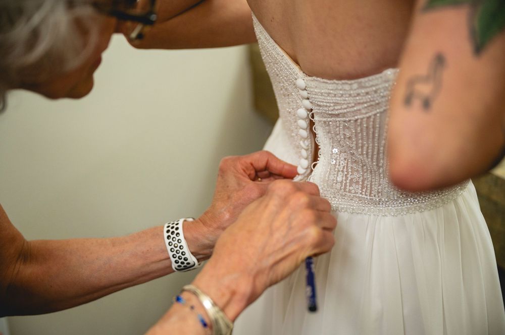 Older woman helping button up the back of a beaded wedding dress on a tattooed bride.