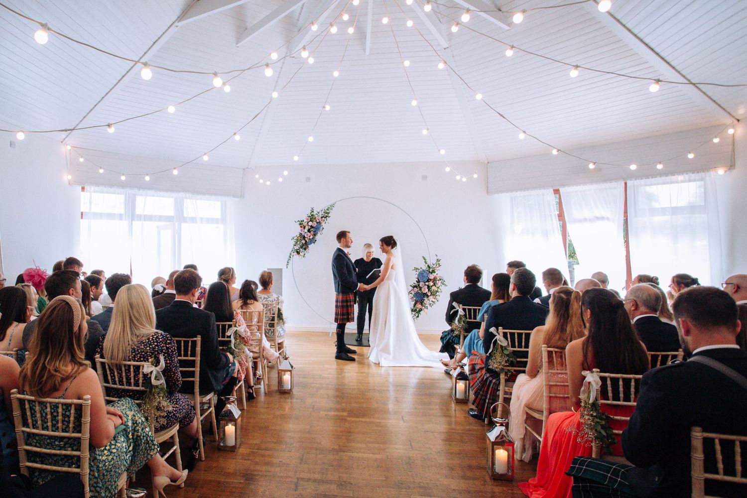 Elegant indoor wedding ceremony under fairy lights with couple at floral arch altar and seated guests on wooden chairs.