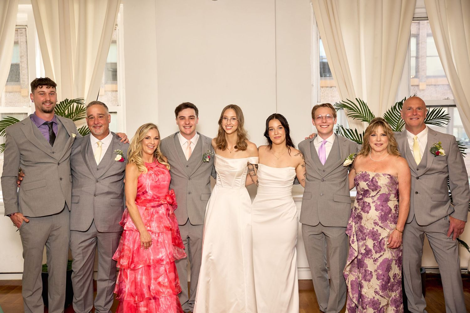 Wedding party posing together in elegant venue, groomsmen in gray suits, bride in white gown, guests in colorful formal attire.