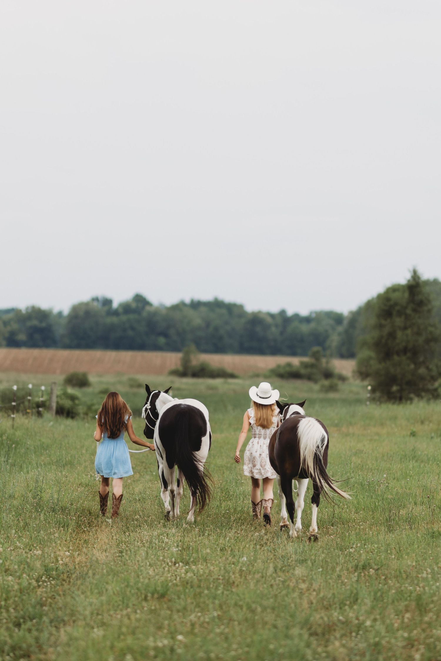 Two girls in cowboy boots and dresses walk with paint horses across a green field on a farm.