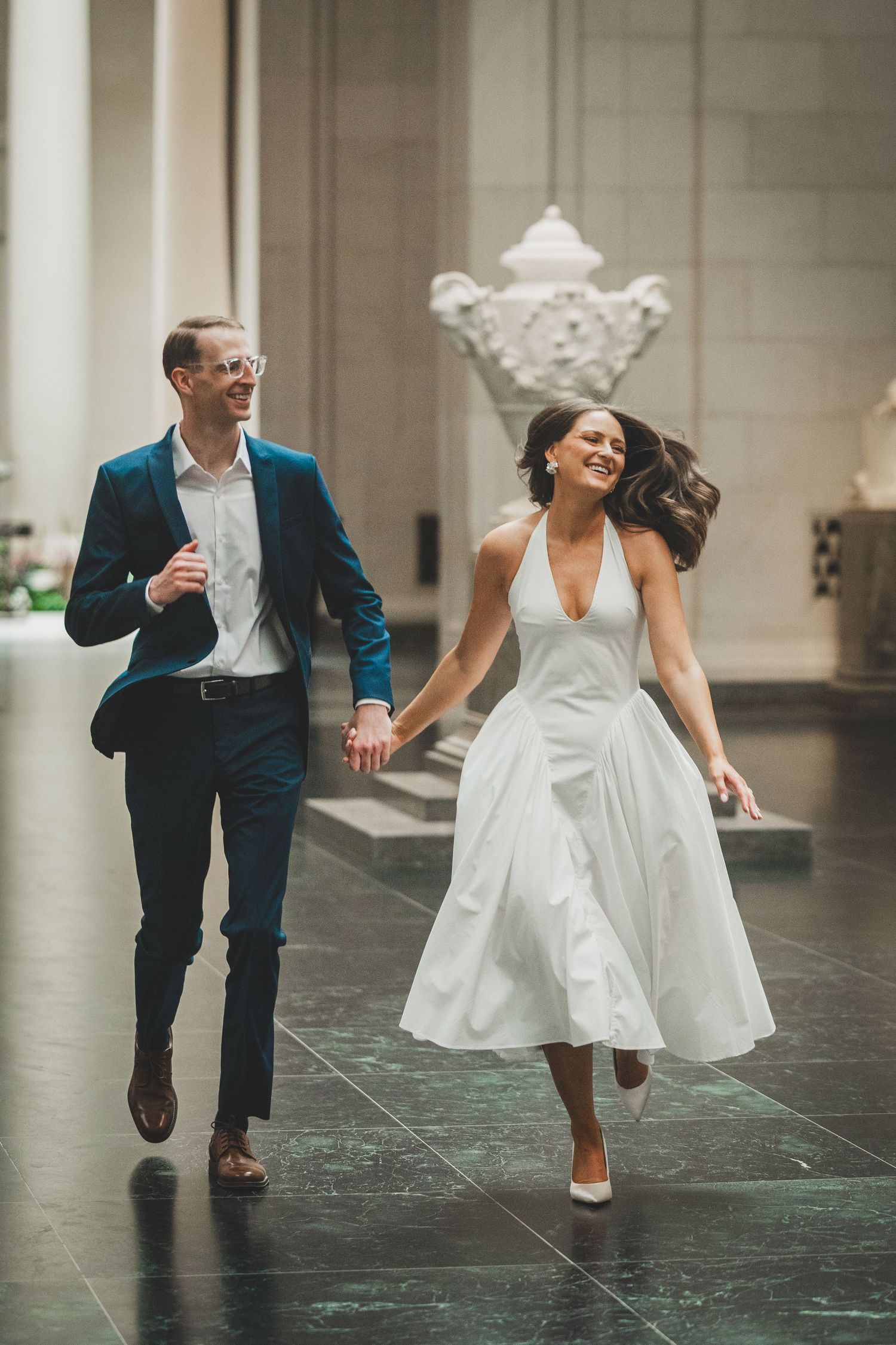A joyful bride in a white halter dress and groom in a teal suit hold hands while running through an elegant hall.