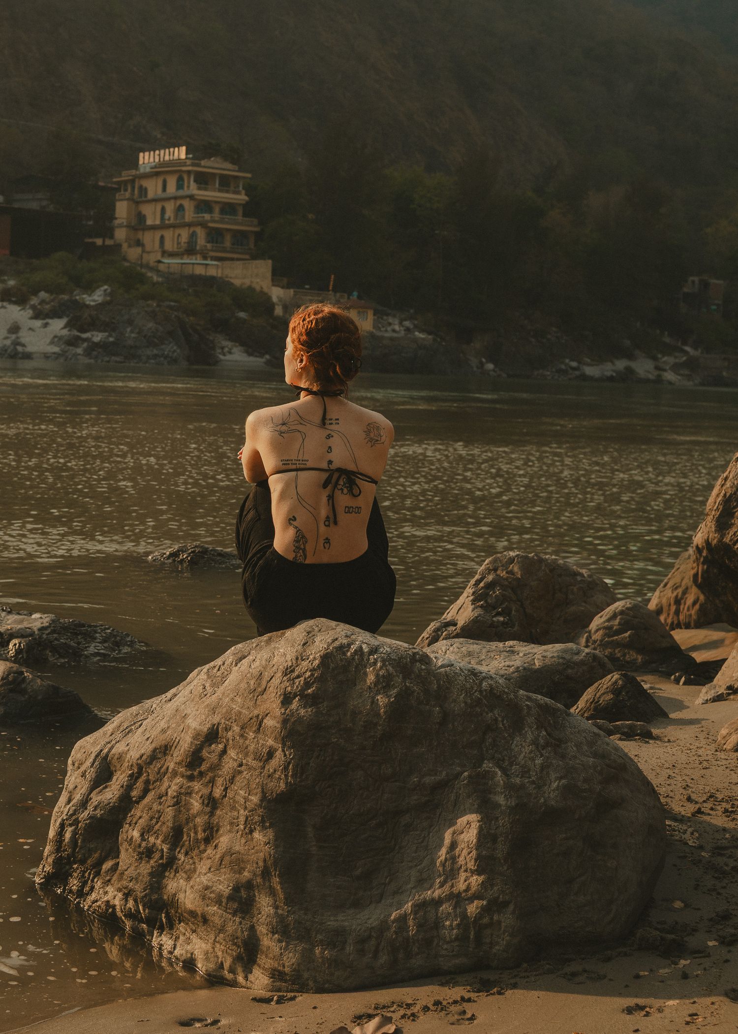 Tattooed woman with red hair sits on rocks by a river, gazing at a hillside building in warm golden light.