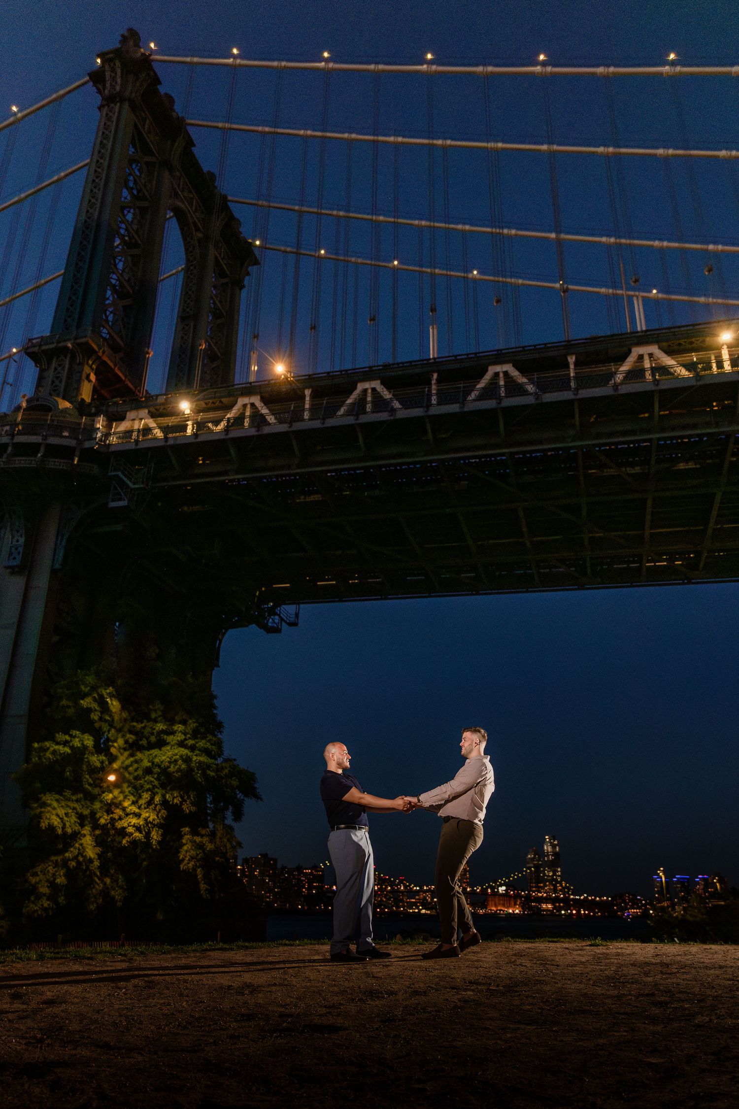 Couple poses on Brooklyn Bridge at dusk with NYC skyline glowing behind them under a blue evening sky.