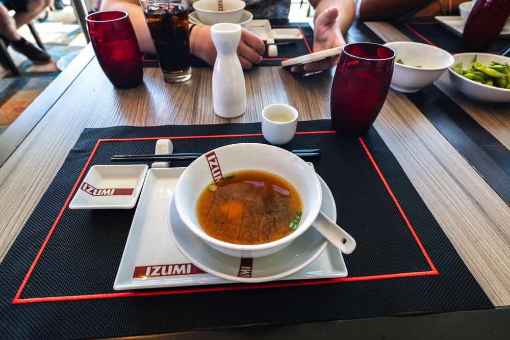 A bowl of miso soup on an Izumi restaurant placemat, with sake and drinks on a dining table.