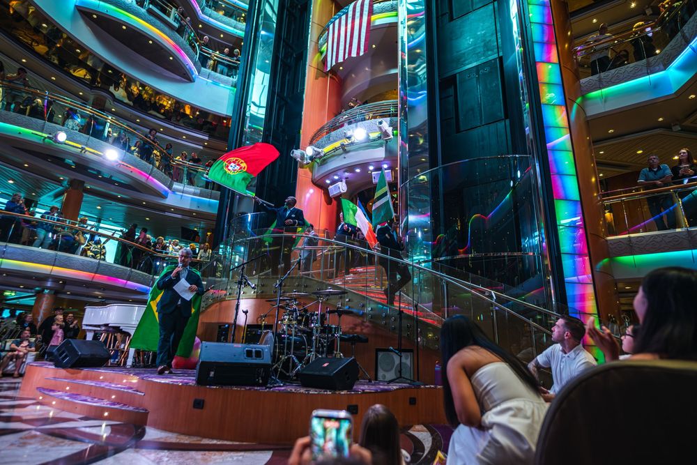 Performers wave colorful flags on a cruise ship atrium stage while guests watch and photograph the lively show.