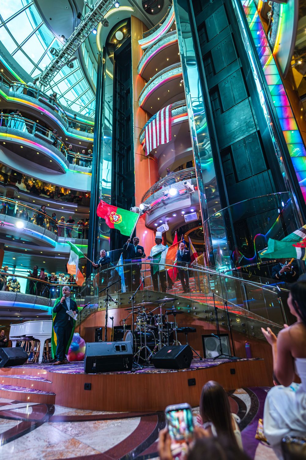 Colorful cruise ship atrium with live band performance, international flags, and guests watching from multiple decks.