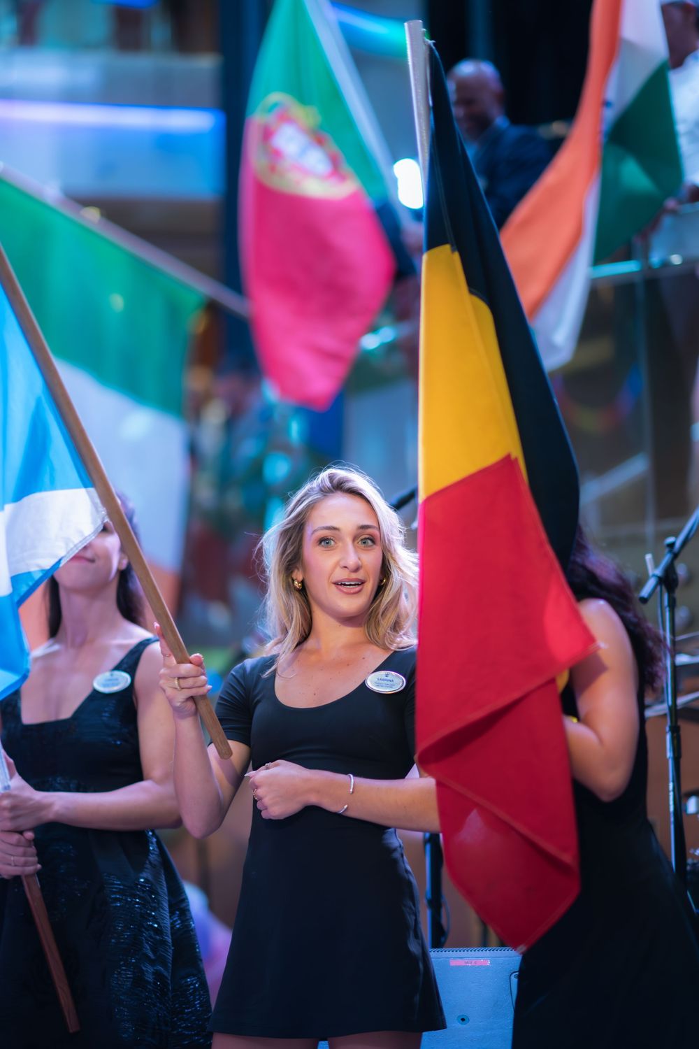 Women in black dresses holding international flags including Belgium and Portugal at a formal ceremony event.