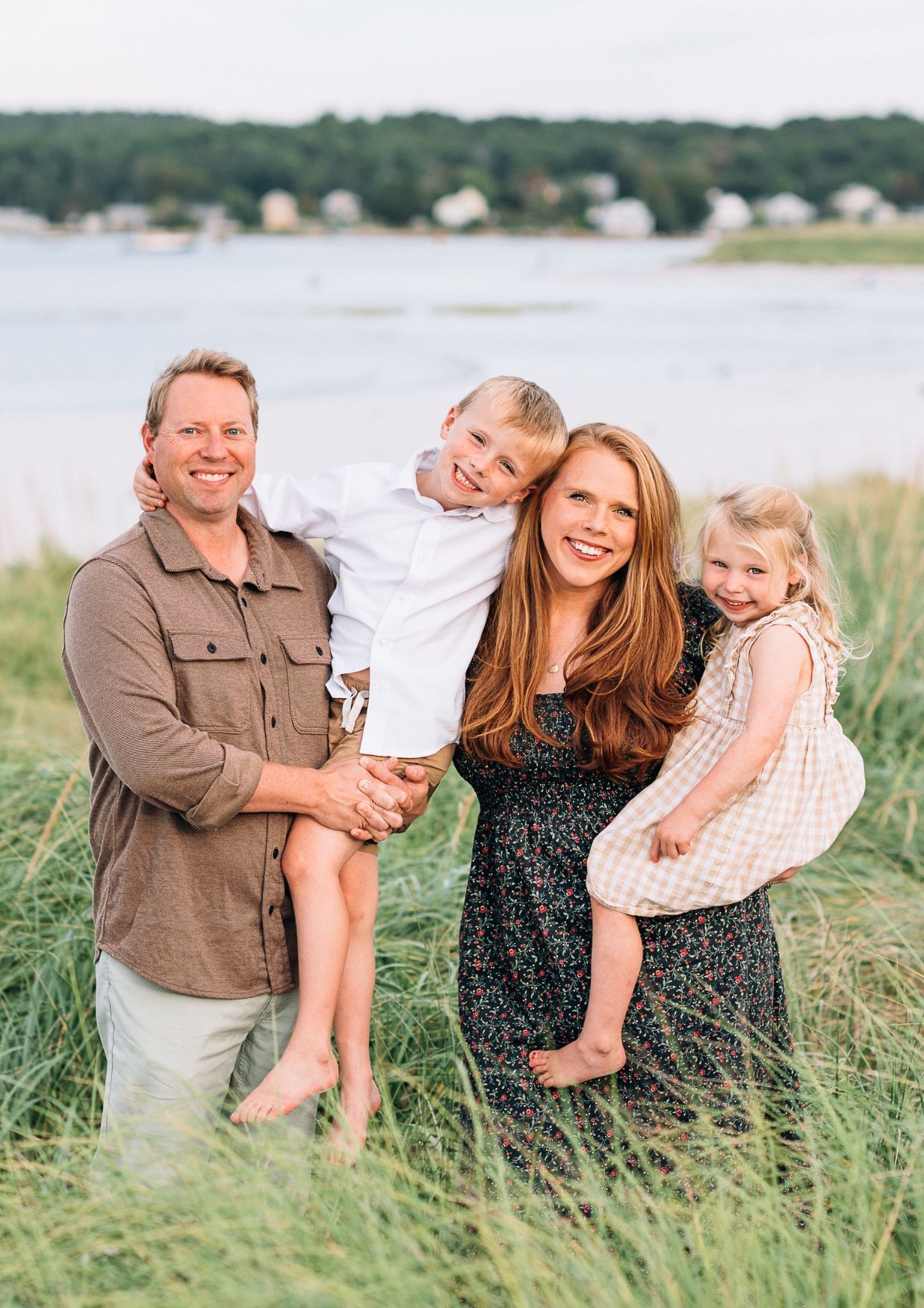 Happy family of four posing outdoors near a coastal waterfront, parents holding two young children in a grassy field.