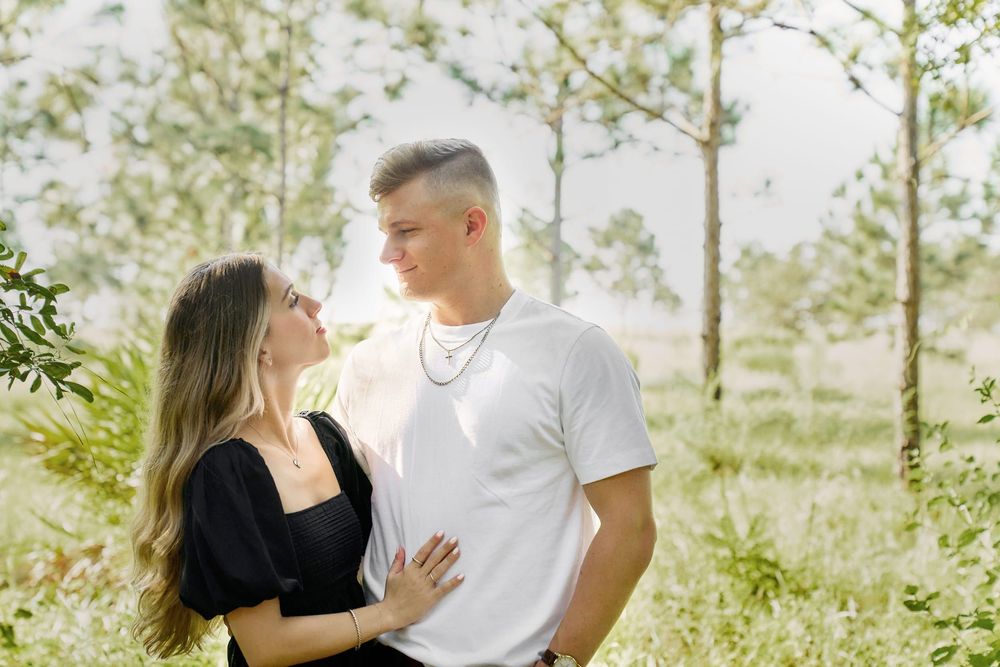 A young couple gazes at each other romantically in a lush green outdoor setting, surrounded by tall trees and wildflowers.