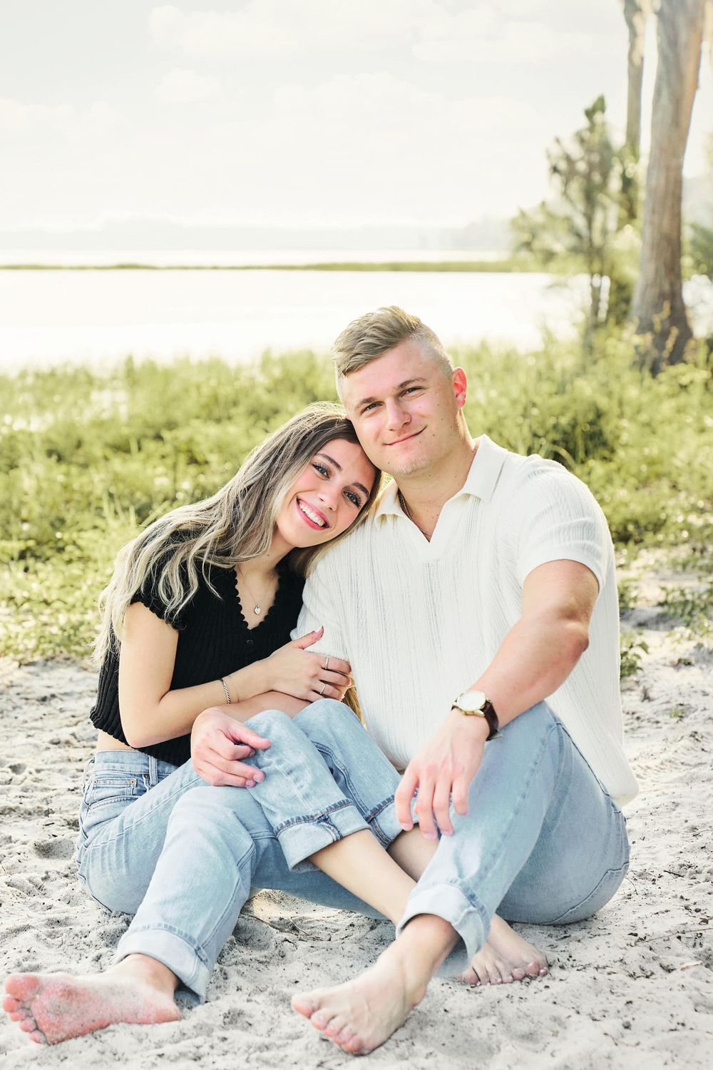 A smiling couple sits barefoot on sandy beach, woman in black top leans on man in white shirt, both wearing jeans.
