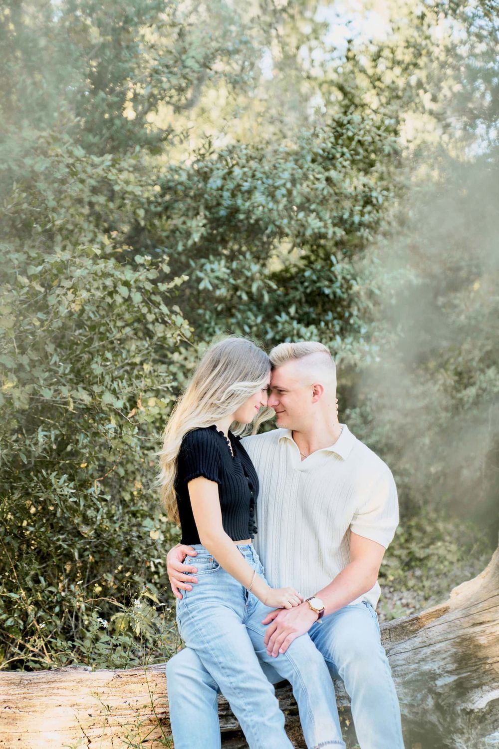 A couple sits on a log, touching foreheads tenderly, surrounded by lush green trees in a romantic outdoor setting.