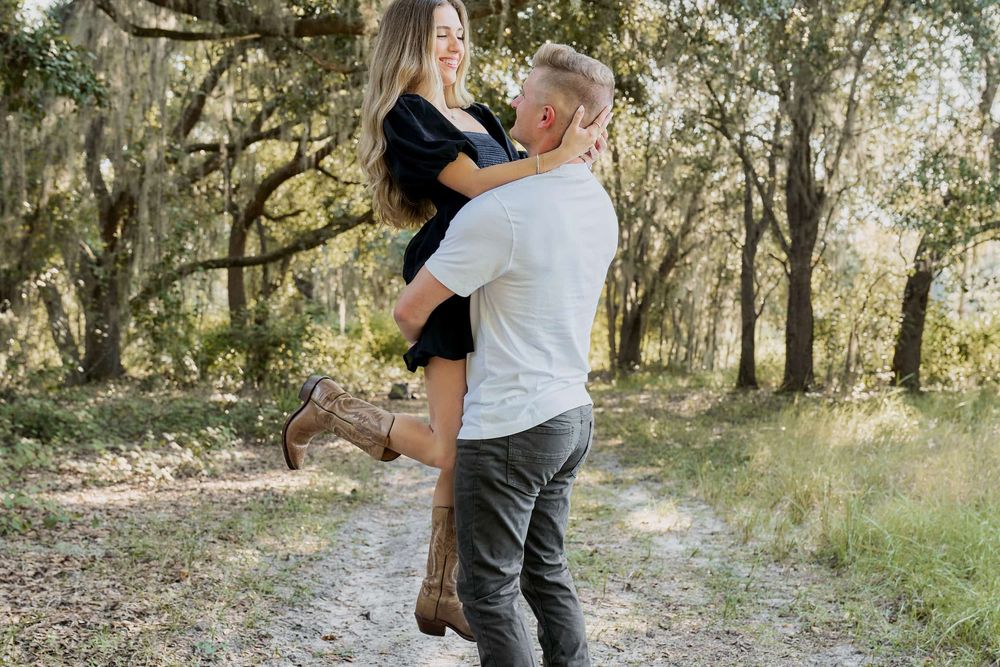 A couple embraces on a wooded path, man lifting woman in black dress and boots among moss-draped trees.