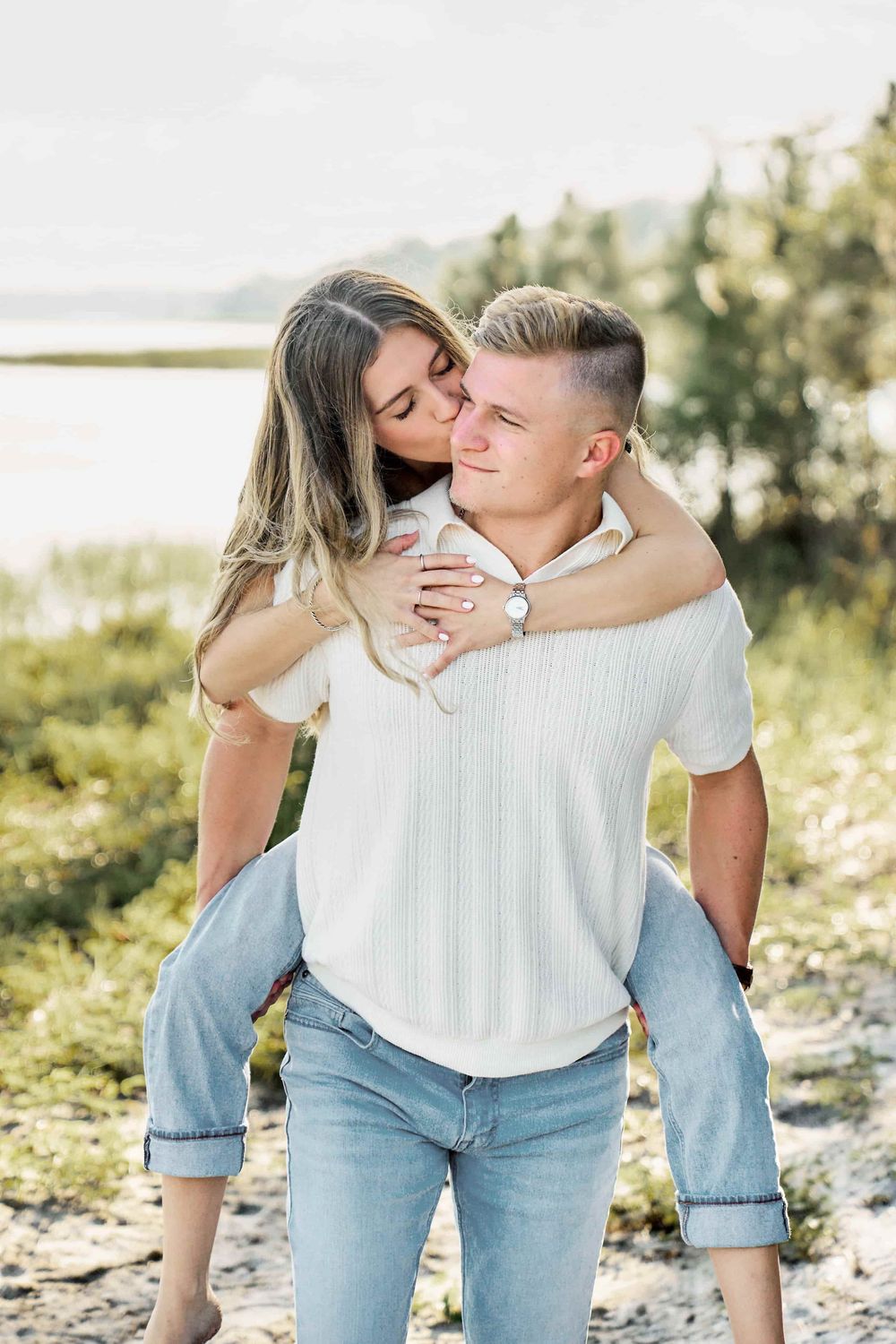 A young couple shares a romantic piggyback ride outdoors near a lake, with the woman hugging the man from behind.