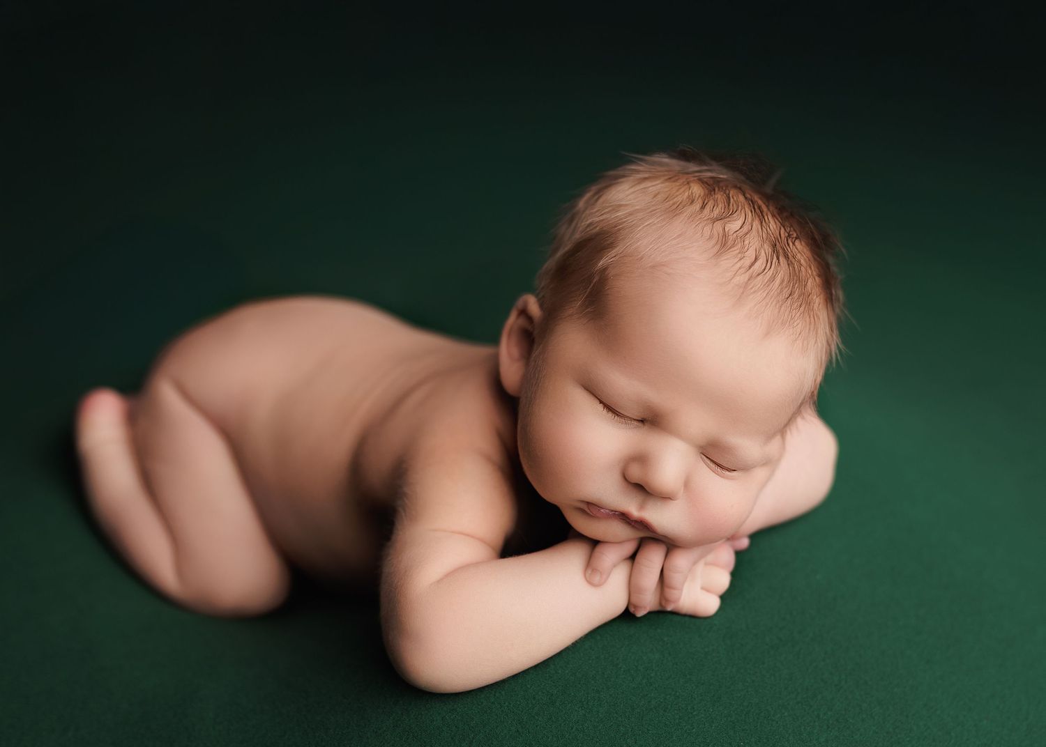 Peaceful newborn baby sleeping on a green backdrop, posed with hands under chin in a classic newborn photography portrait.