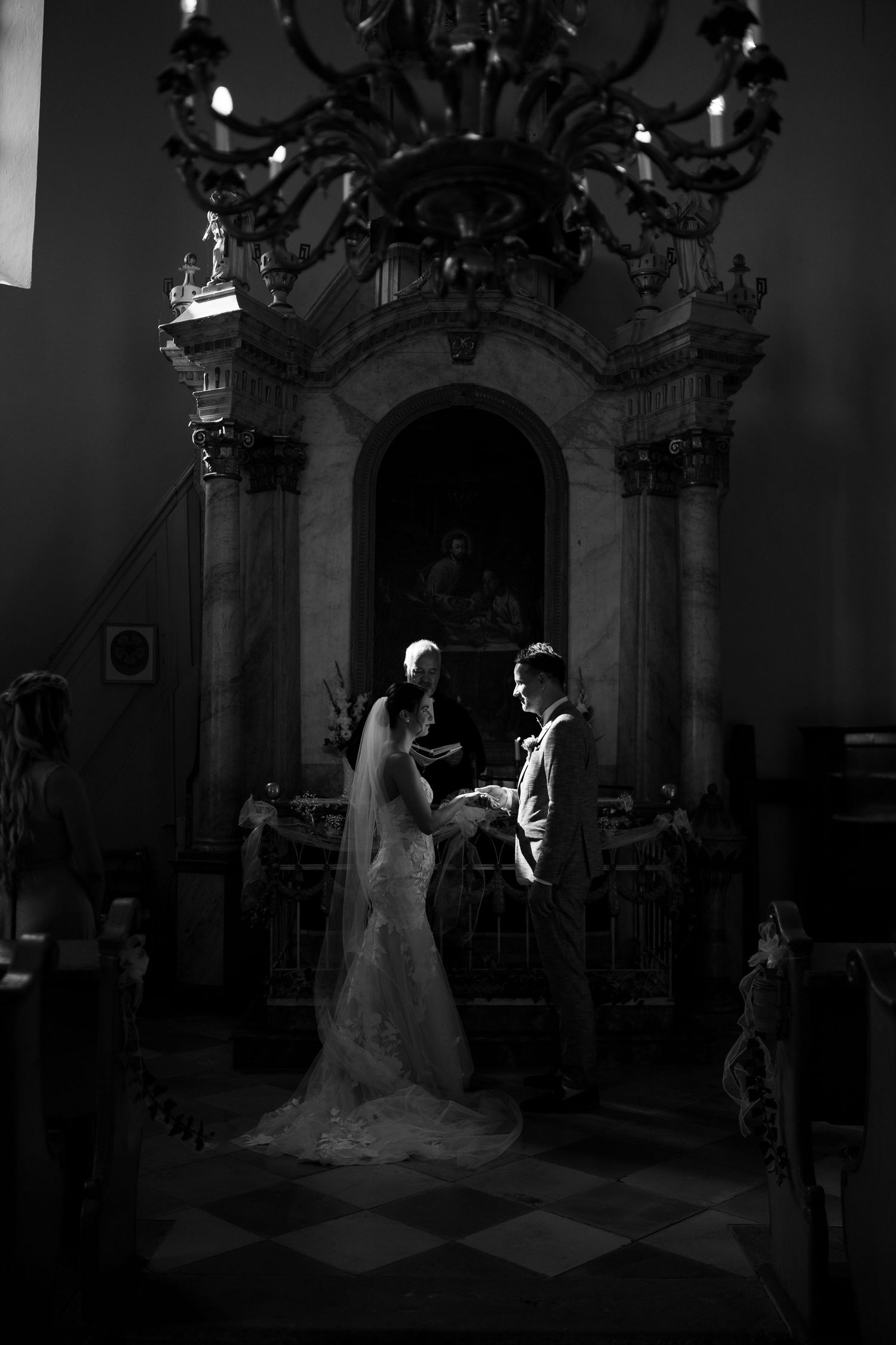 Black and white photo of bride and groom exchanging vows at ornate church altar with chandelier overhead.