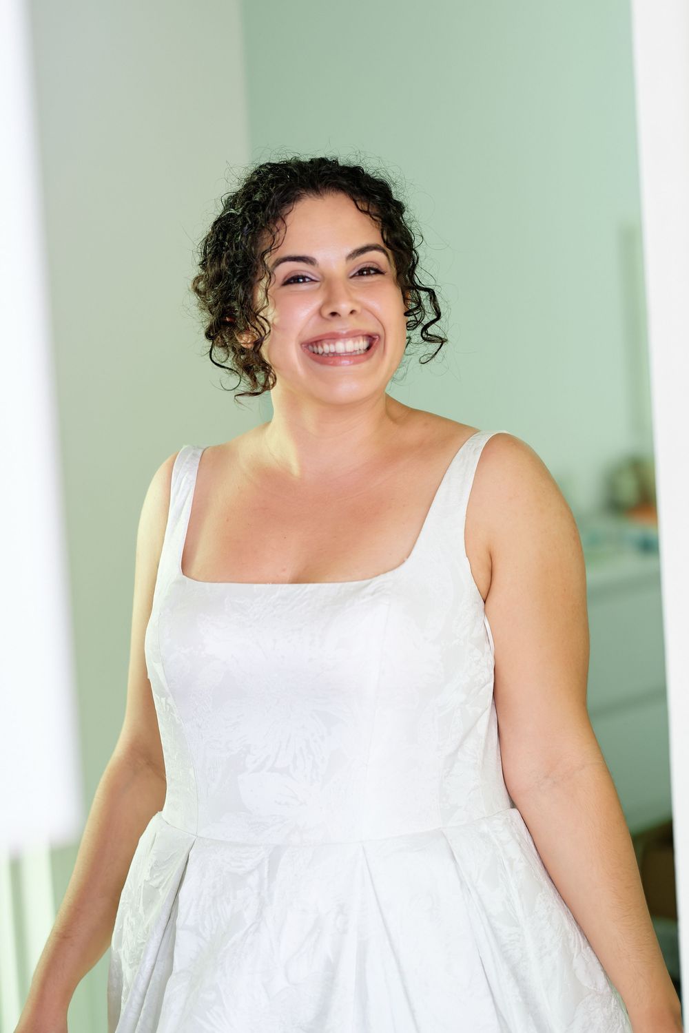 Smiling bride in white sleeveless wedding dress with curly dark hair posing indoors.