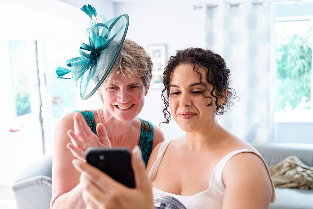 Two women smile while viewing photos on a smartphone, one wearing a teal fascinator hat at a wedding.