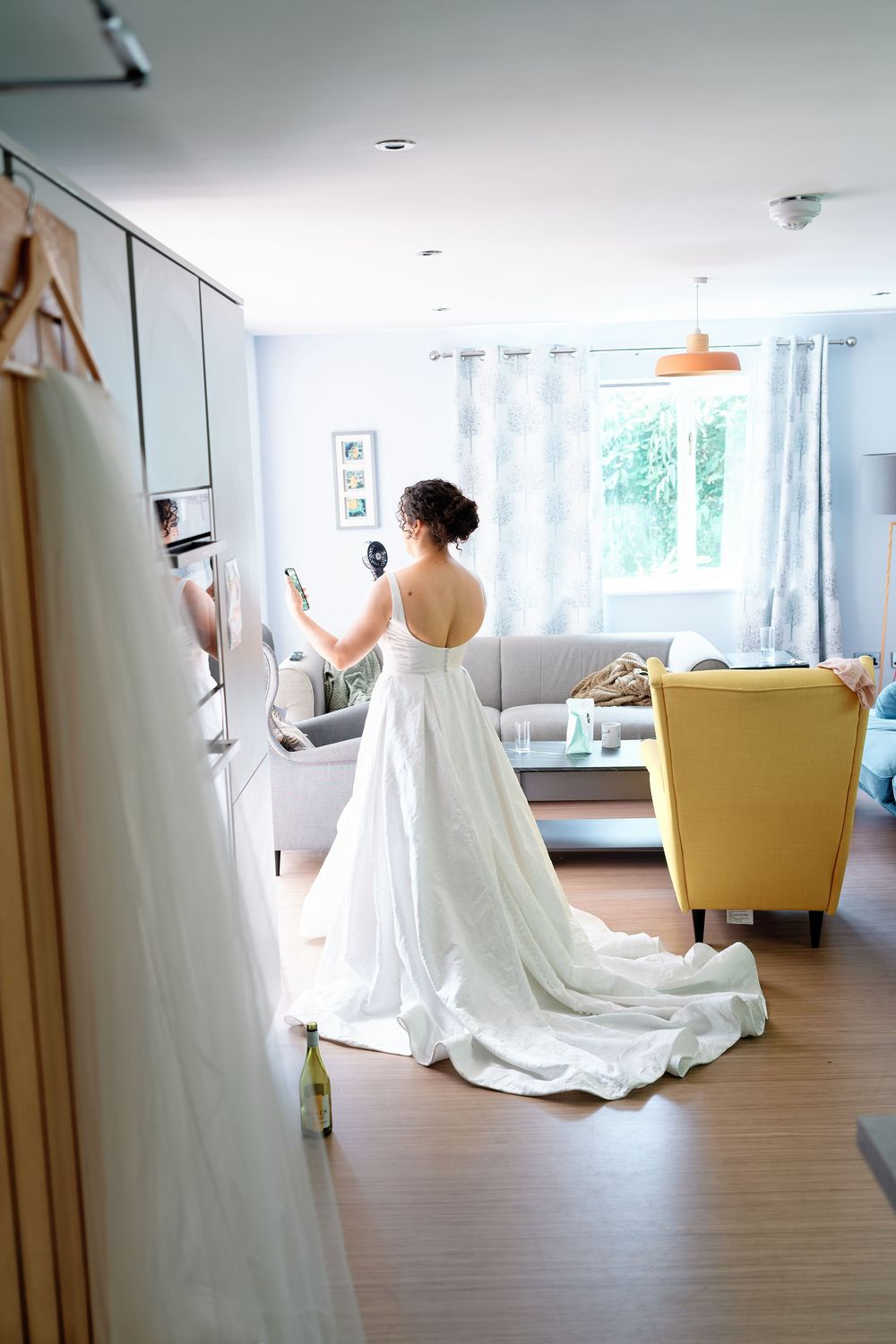 Bride in a stunning white backless wedding gown with a long train standing in a bright modern living room.