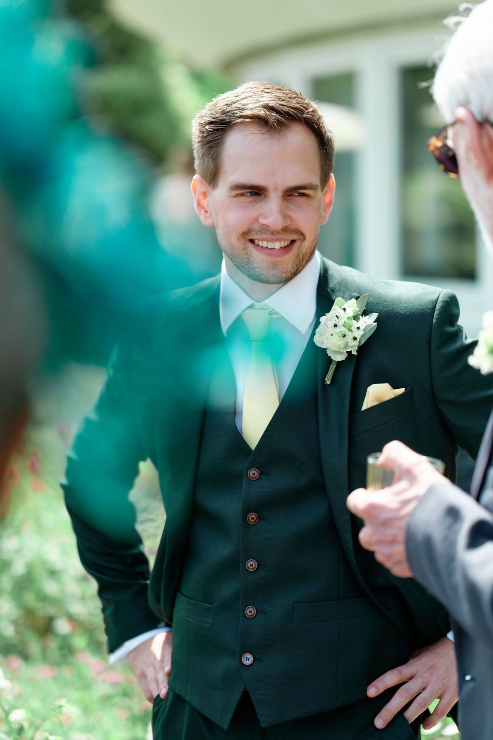 Smiling groom in dark green suit with boutonniere and yellow tie at outdoor wedding ceremony.