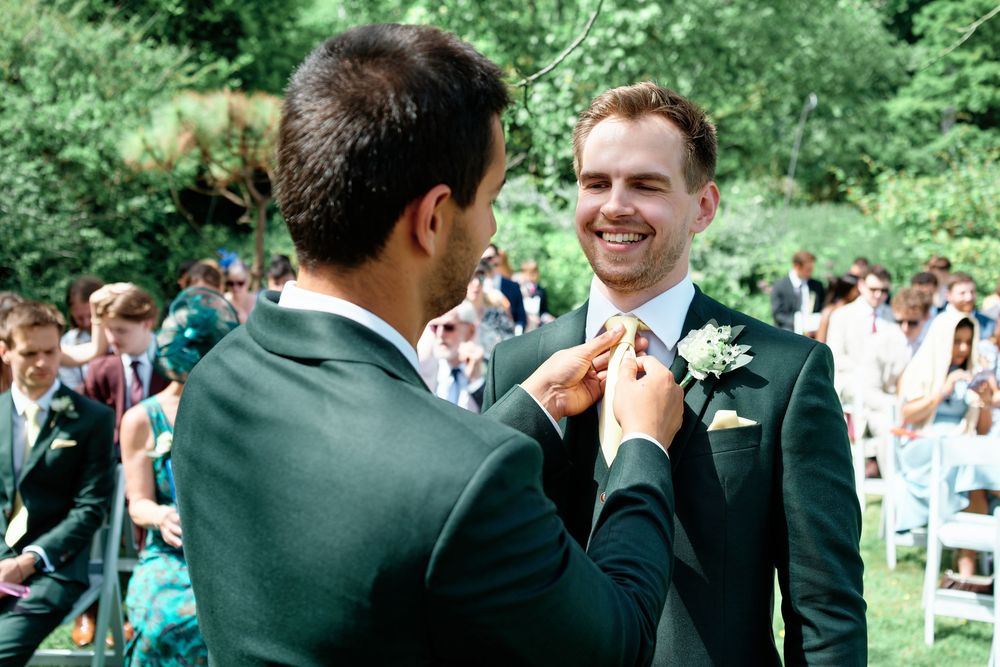 Groom smiles as groomsman adjusts his boutonniere at an outdoor wedding ceremony.