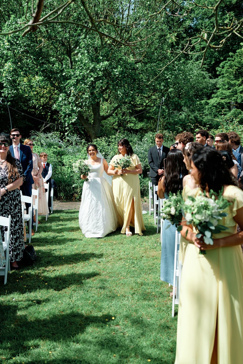 Bride walks down outdoor aisle with bridesmaid in yellow dress, surrounded by guests and lush greenery.