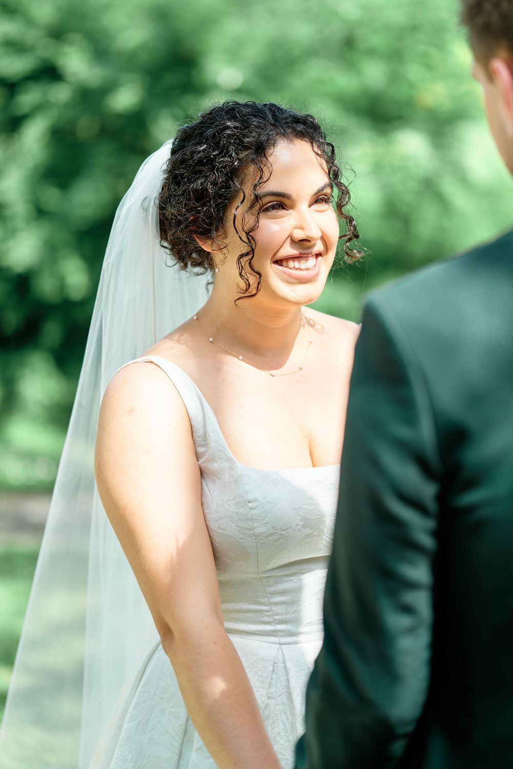 Smiling bride in white gown and veil stands outdoors facing groom during wedding ceremony.