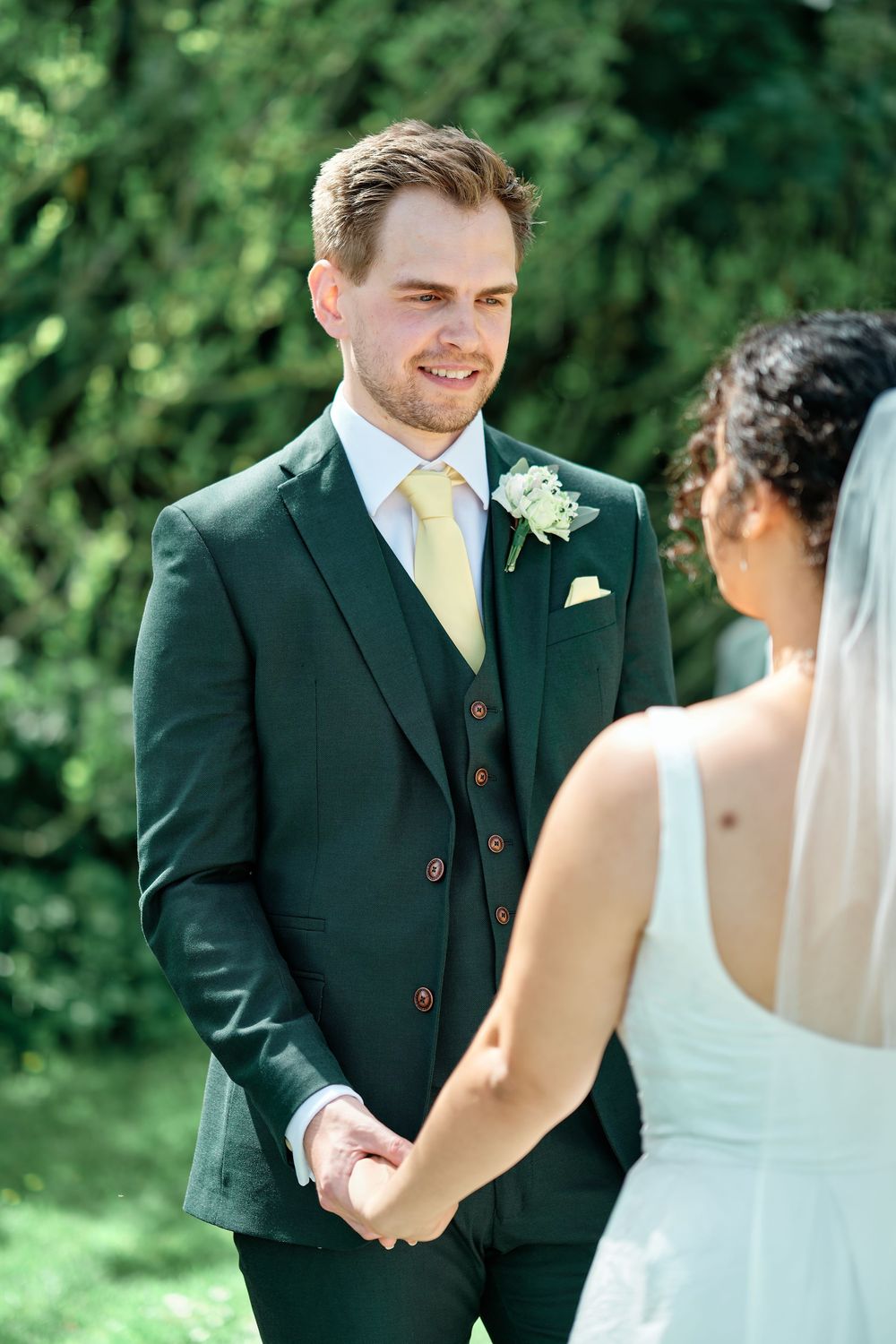 Groom in dark green three-piece suit holding hands with bride outdoors during wedding ceremony.