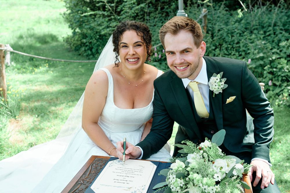 Happy newlyweds signing marriage certificate outdoors, bride in white gown with veil, groom in dark suit with yellow tie.