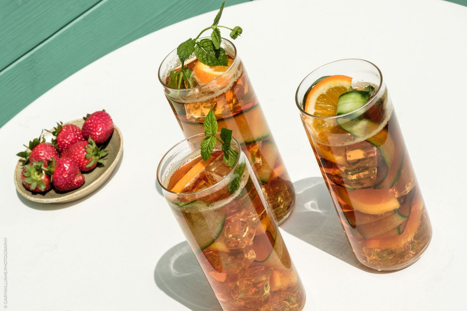 Three tall glasses of Pimm's cup cocktail with fruit and mint on a white table beside fresh strawberries.