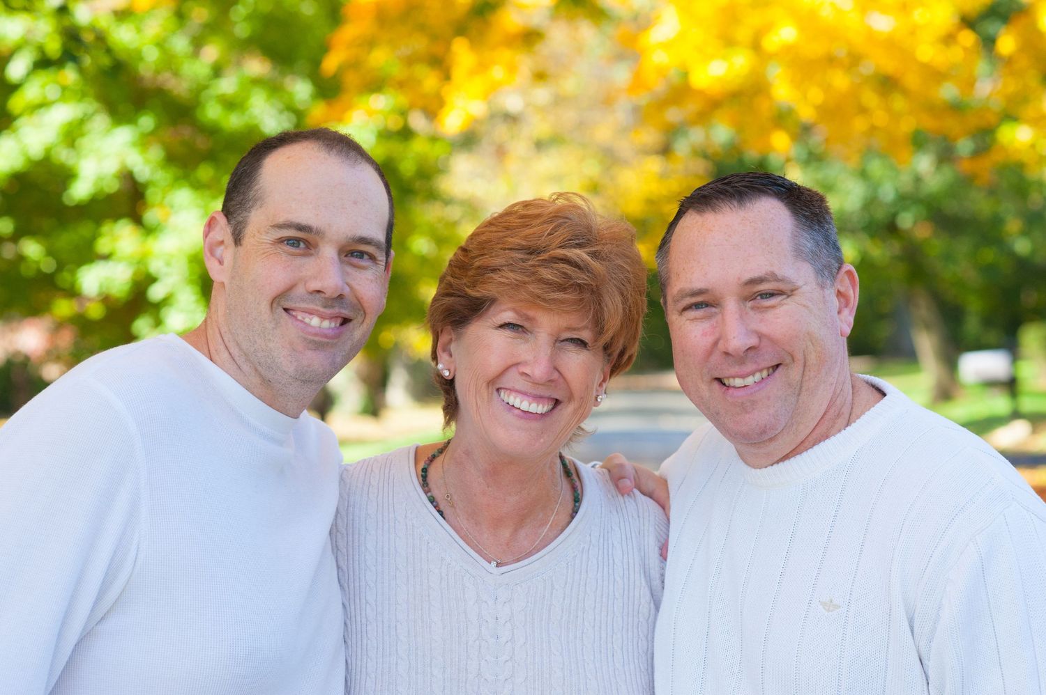 A smiling woman with short red hair poses outdoors with two men in white shirts, framed by vibrant autumn foliage.