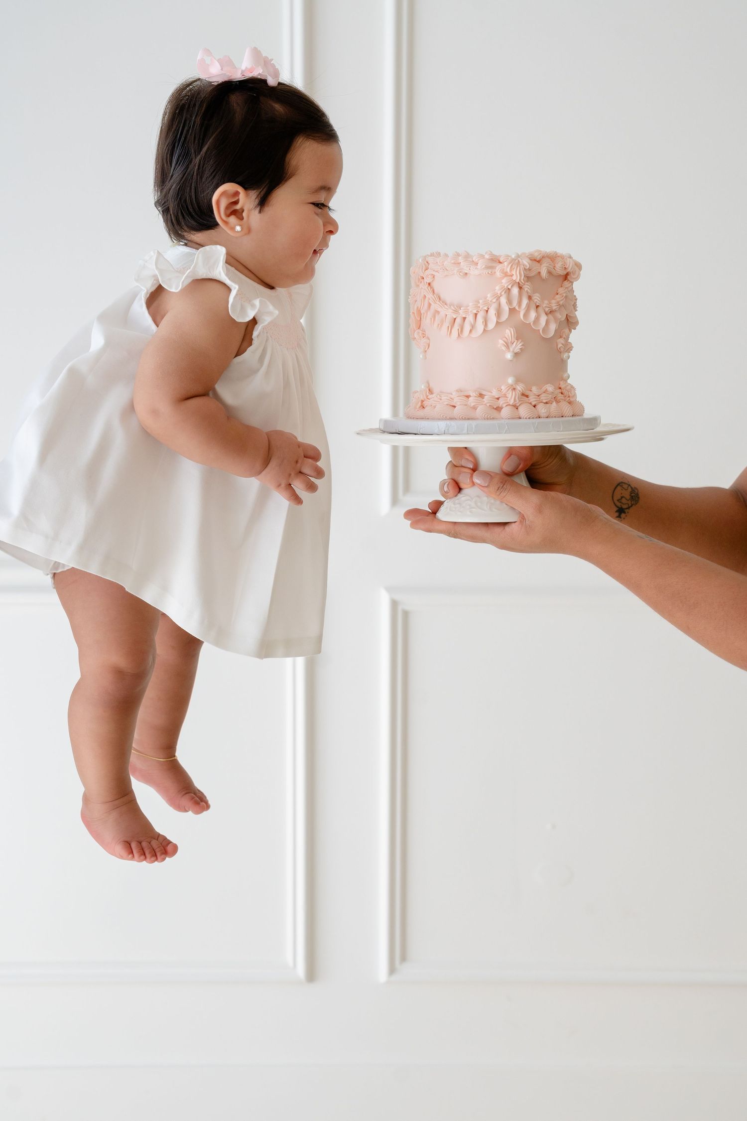 A baby girl in a white dress reaches toward a pink birthday smash cake held on a stand against a white wall.
