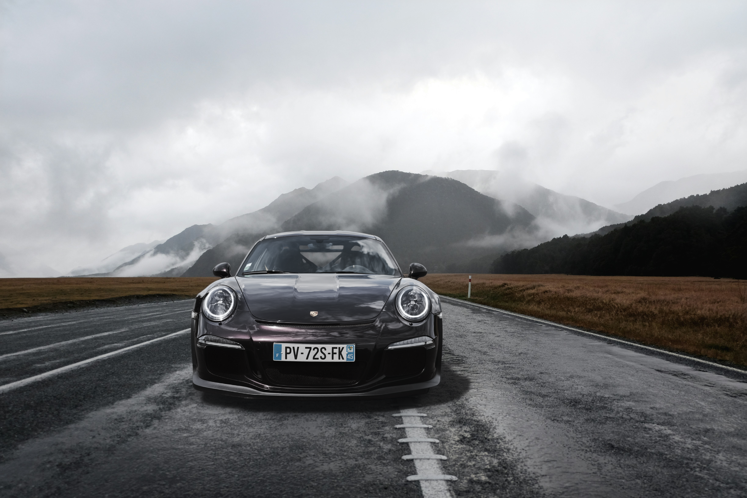 Dark grey Porsche 911 GT3 RS parked on misty mountain road with dramatic cloudy landscape backdrop.