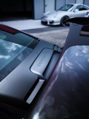Close-up of a dark Porsche rear spoiler with embossed logo, white Porsche 911 visible in background.