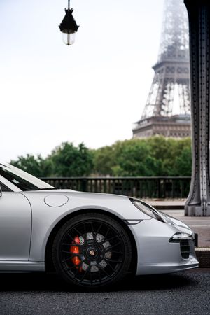 White Porsche 911 parked near the Eiffel Tower in Paris, France, showcasing its sleek design and black alloy wheels.