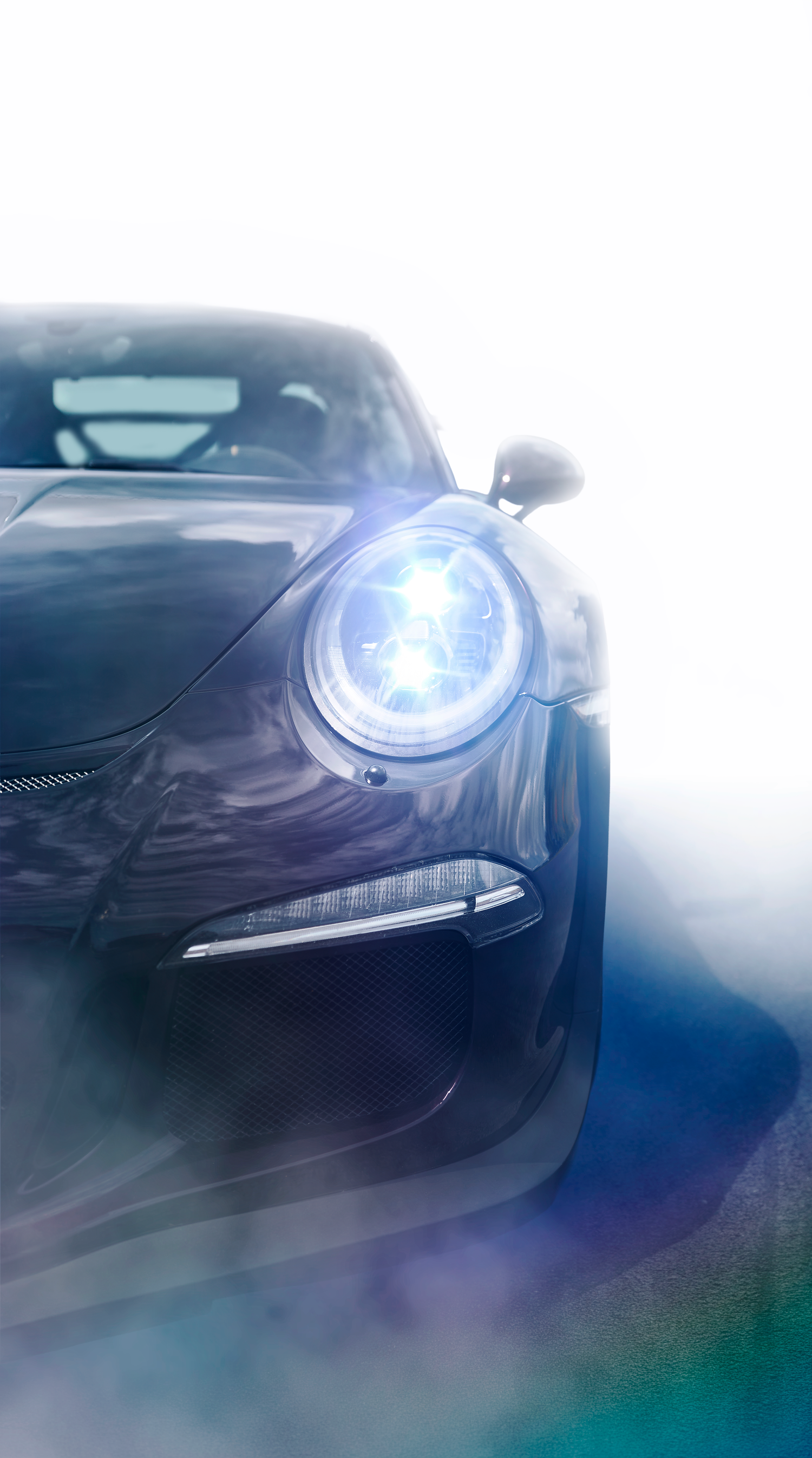 Dark Porsche 911 GT3 sports car front view with glowing blue HID headlight emerging from white misty background.